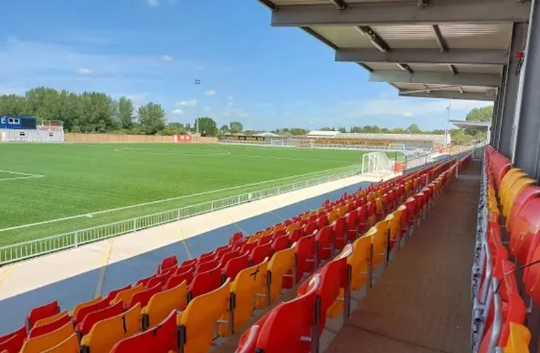 Góc nhìn từ khán đài sân Meadow Park, bao quát toàn bộ mặt sân cỏ nhân tạo và một phần khán đài đối diện trong ánh nắng chiều.
