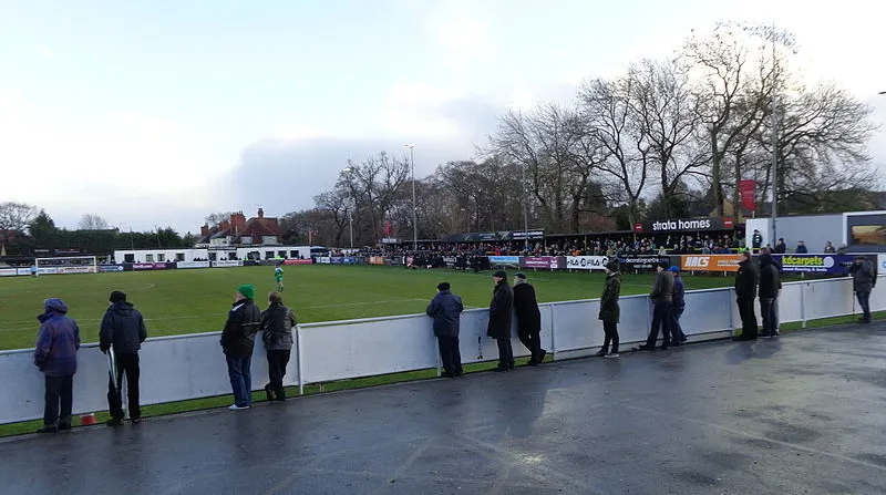 Khoảnh khắc trận đấu giữa Harrogate Town và Bradford Park Avenue diễn ra tại sân Wetherby Road, với các cầu thủ đang tranh bóng trên nền khán đài đơn giản, thể hiện tinh thần thi đấu của một câu lạc bộ hạng dưới.